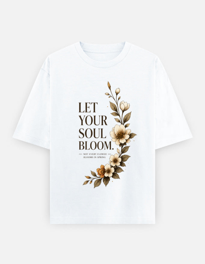 A white short-sleeve crewneck t-shirt featuring a graphic print on the front with the text 'LET YOUR SOUL BLOOM.' in a dark brown serif font, accompanied by a vertical floral illustration in shades of beige, brown, and muted green.