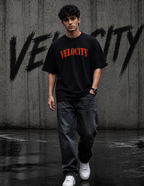 A man wearing a black t-shirt with 'VELOCITY' printed in orange across the chest, paired with dark gray jeans and white sneakers, walking in front of a concrete wall with graffiti.